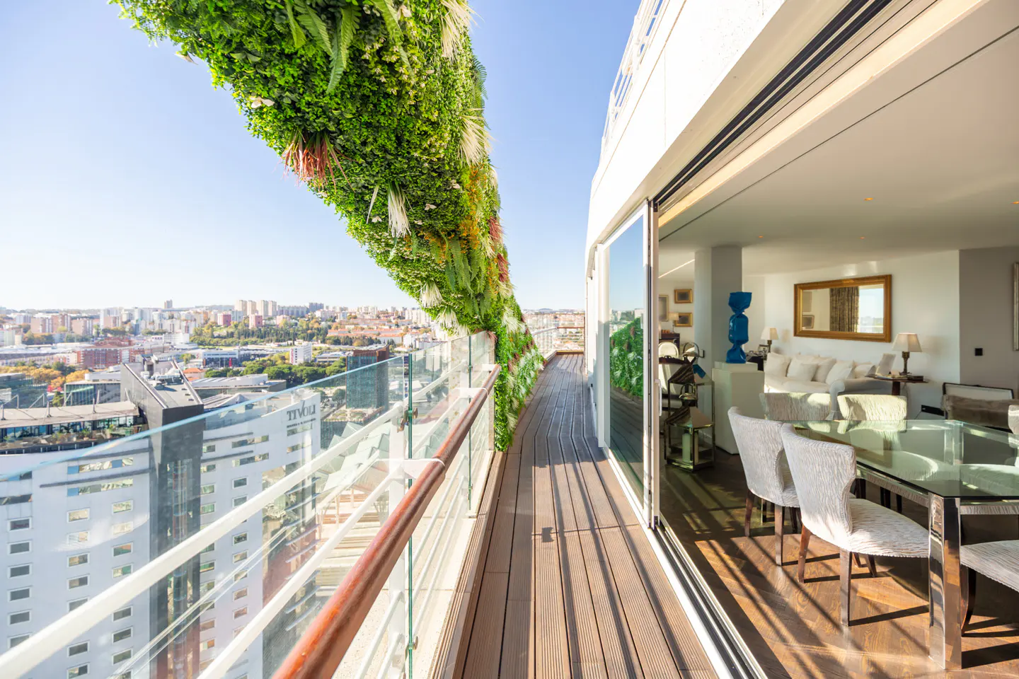 Bright apartment balcony with a green wall, glass railing, and wood floor overlooking a city. Sliding glass doors open to a living room with a glass table and white furniture.