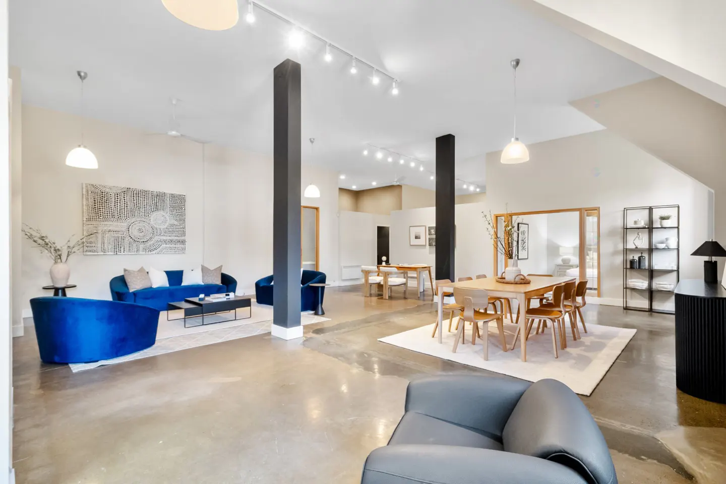 Bright, open-concept living space with blue velvet sofas, wood dining table, and polished concrete floors. Black support columns add architectural interest.