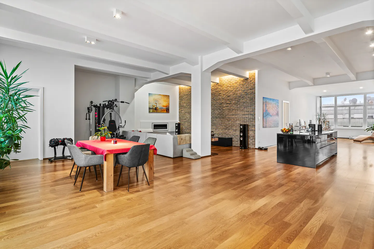Open-concept loft with hardwood floors, white walls, and exposed beams. A dining table, gym equipment, and kitchen island are visible.