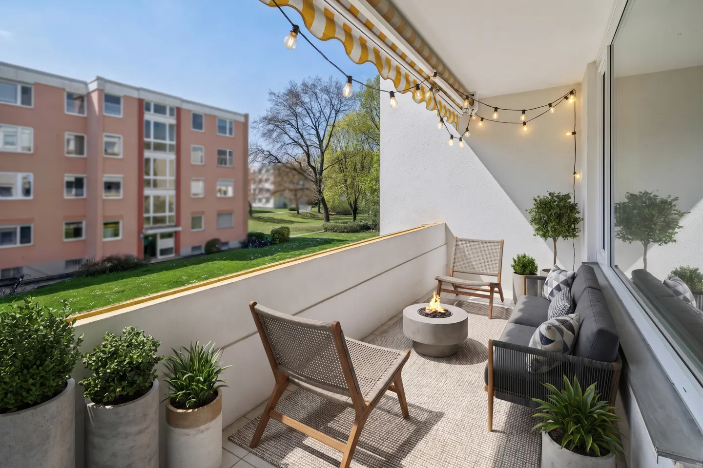 A bright balcony with a yellow awning, string lights, and outdoor furniture including chairs, a sofa, and a small fire pit. Green plants add a touch of nature.