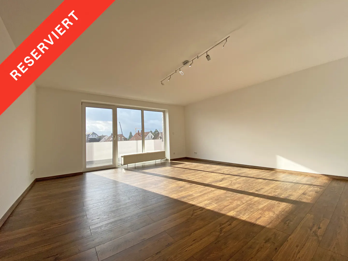 Bright, empty room with wood floors, white walls, and a sliding glass door to a balcony. A red banner in the corner says "Reserviert".