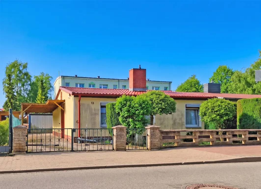 A one-story yellow house with a red roof and chimney, a black iron gate, and green bushes under a clear blue sky.