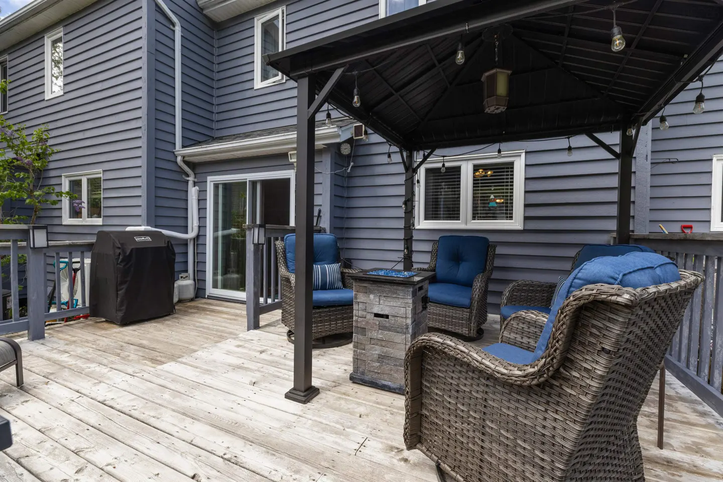 Back deck with a gazebo, wicker chairs with blue cushions, a stone fire pit, and a grill against a grey-sided house.