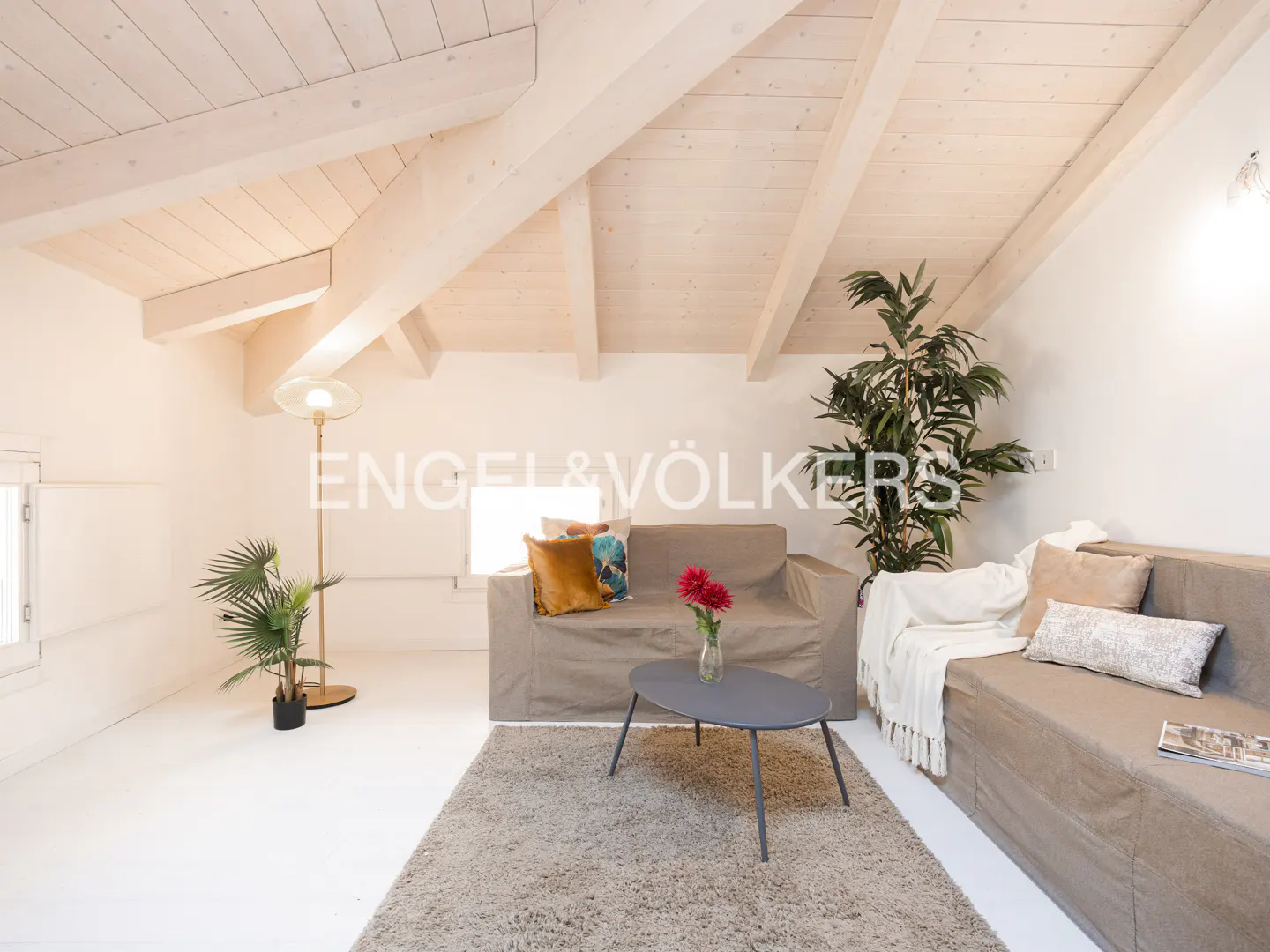 Attic living room with beige sofas, a grey coffee table, a rug, and potted plants under a white wooden beamed ceiling.
