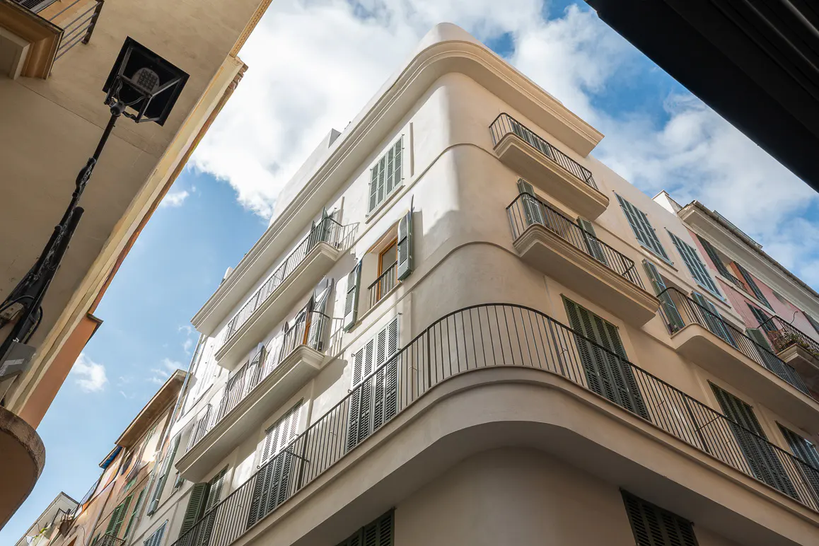 Low angle view of a white building with balconies and green shutters against a blue sky.