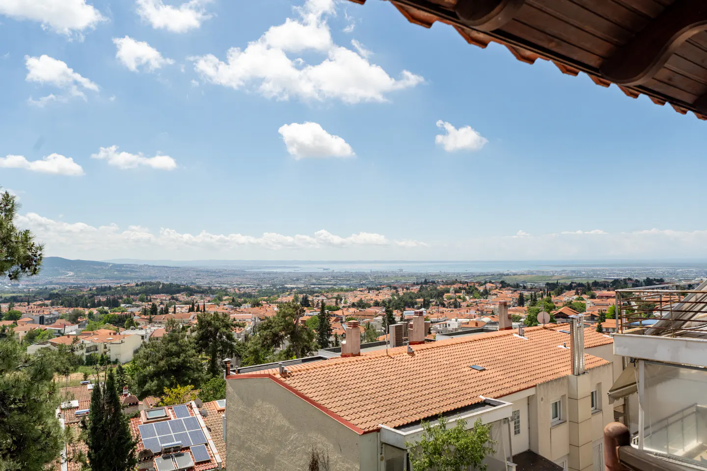 View from a balcony overlooking a city with red-tiled roofs, green trees, and a distant body of water under a blue sky with white clouds.