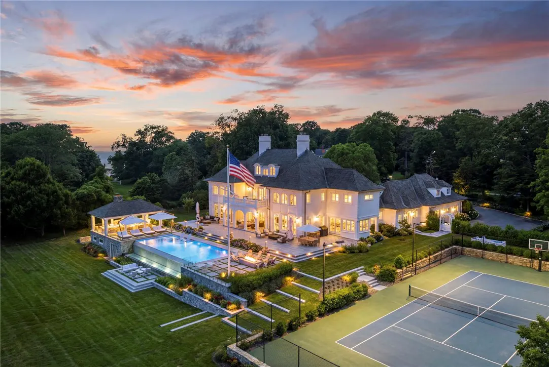 Aerial view of a large white house with a pool, tennis court, and American flag at sunset.