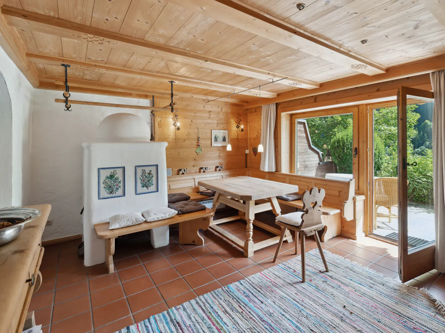 Rustic dining room with wood-paneled walls, a built-in bench, a wooden table, and a white tiled stove. A colorful rug lies on the terracotta tile floor.