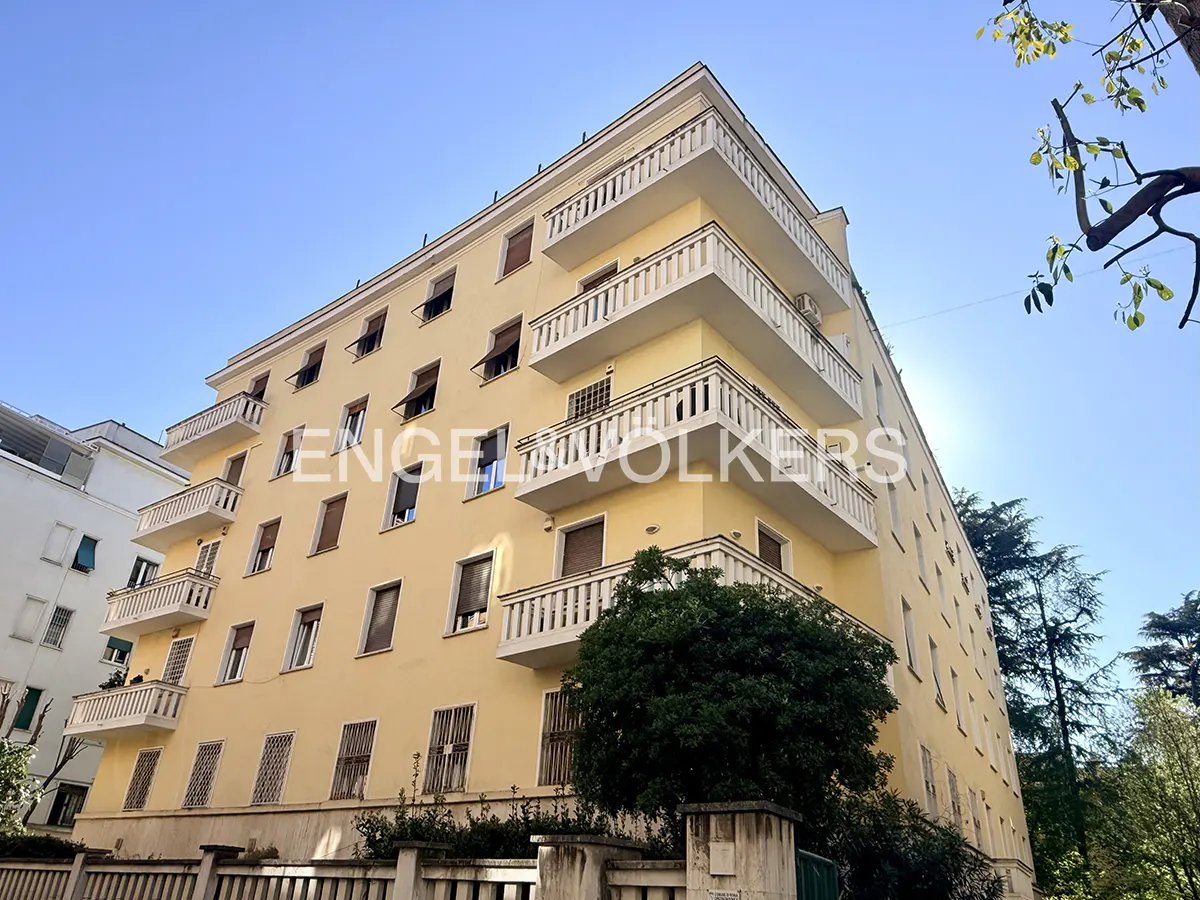 Exterior view of a yellow apartment building with white balconies under a blue sky. Trees surround the building.