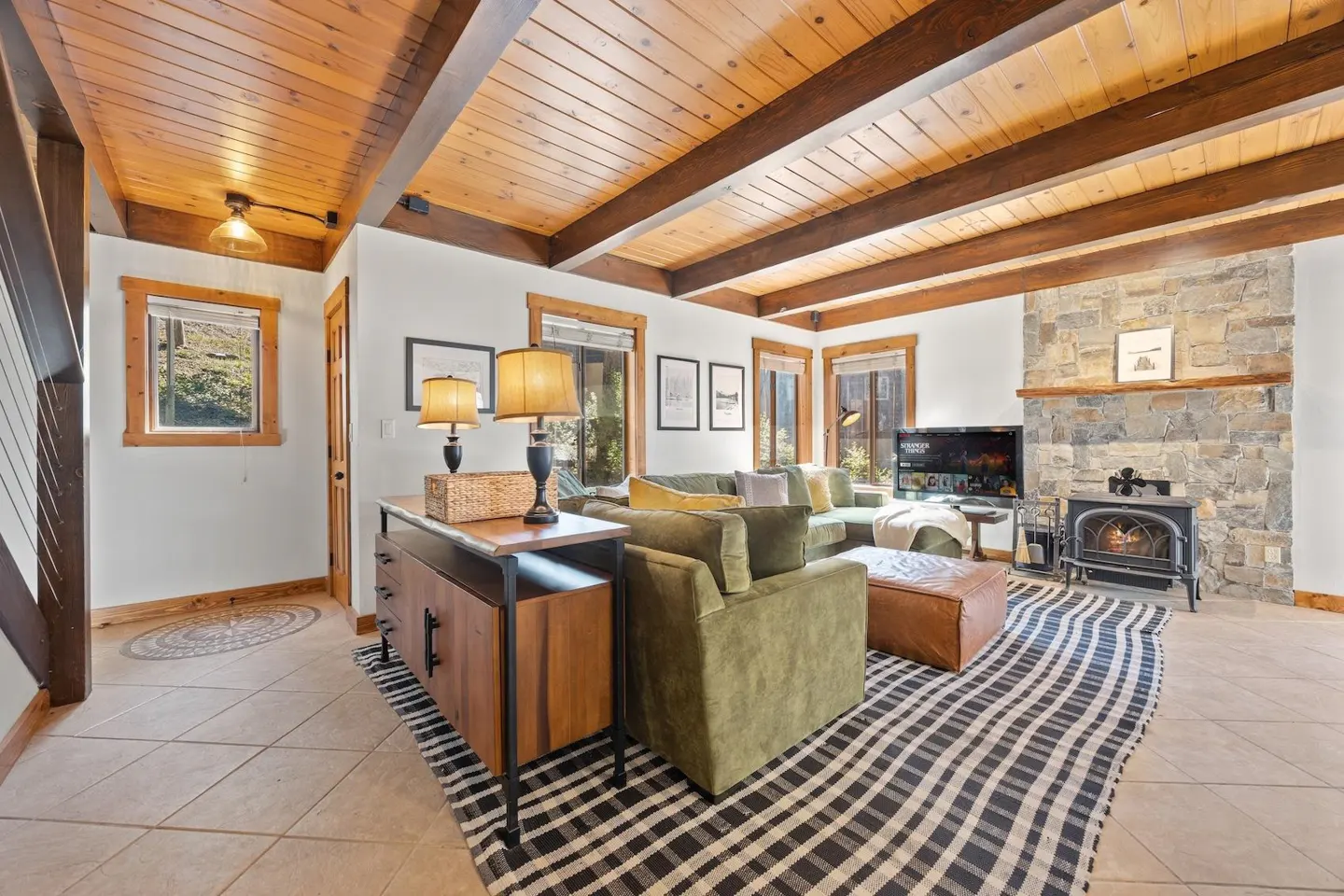 Living room with wood ceiling beams, stone fireplace, and green sofa. A black and white rug sits on the tile floor.
