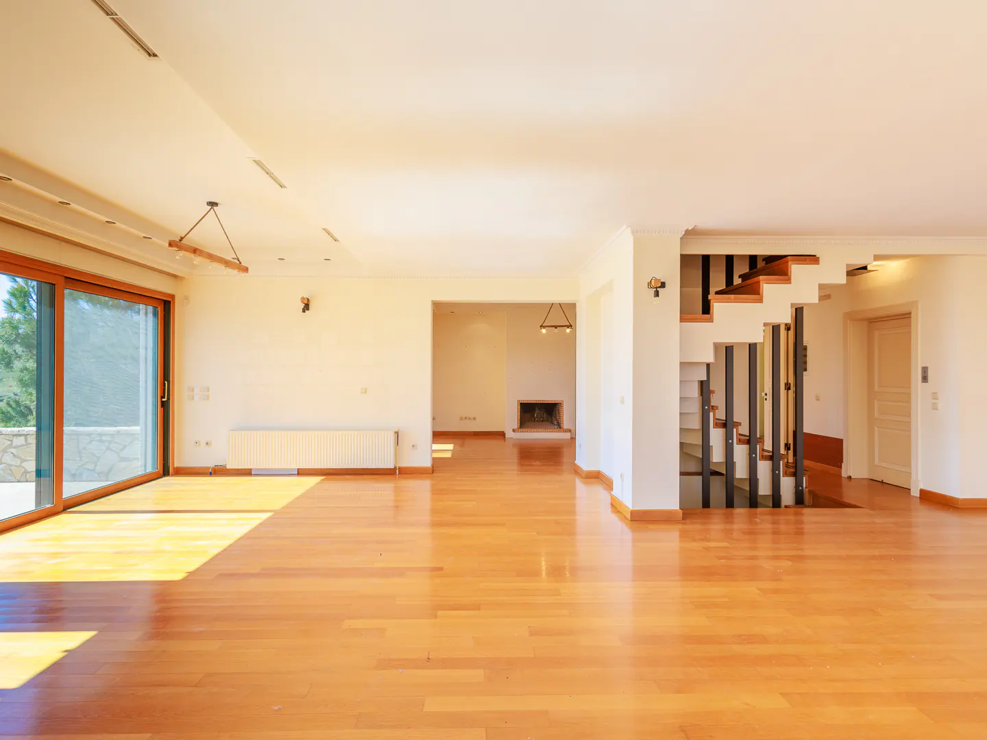Bright, empty living room with hardwood floors, white walls, and a staircase. A fireplace is visible in the background. Sliding glass doors to the left.