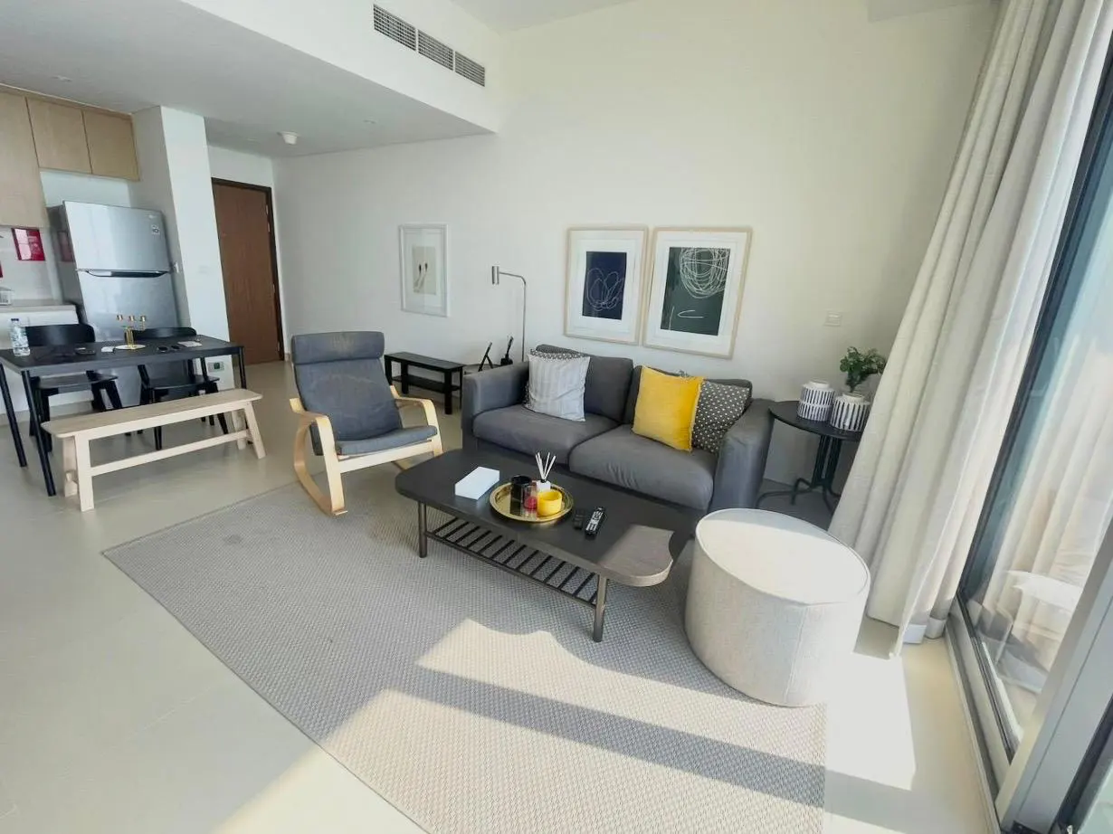 Bright apartment living room with gray sofa, rocking chair, coffee table, and dining area. Large windows with white curtains.
