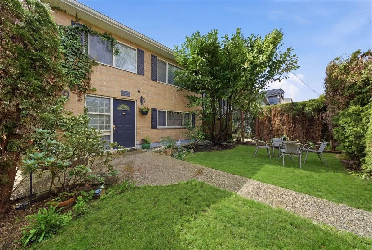 Two-story tan brick building with a blue door, black shutters, and a green lawn with a patio set.