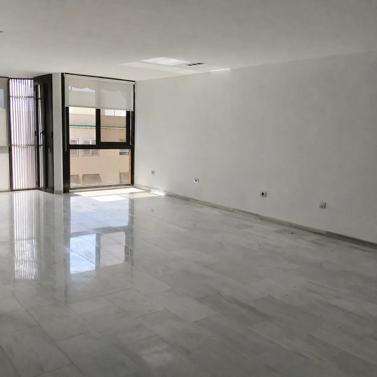 Empty room with white walls and a marble floor. A large window with a roller blind is on the left.