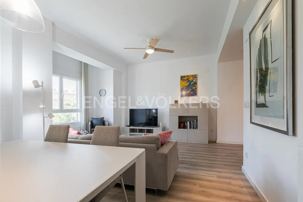 Bright, modern living room with a white table, gray sofa, TV, and light wood floors. A ceiling fan and artwork add detail.