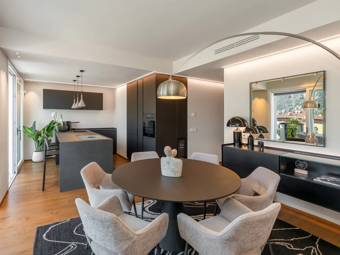 A modern apartment interior with a black round table surrounded by six gray chairs, a black kitchen, and a large mirror.