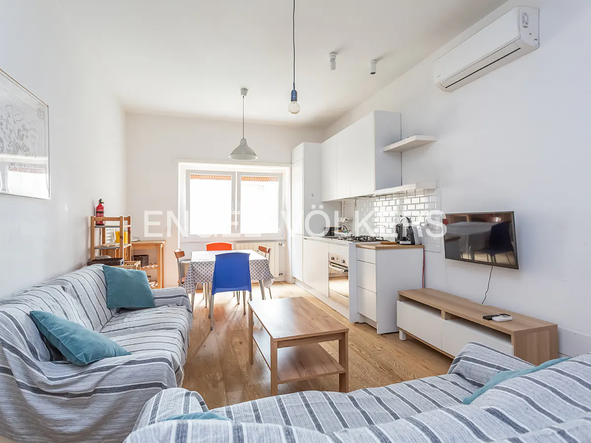 Bright, open-concept apartment with striped sofas, a wooden coffee table, and a dining area near a window. A white kitchen is visible in the background.
