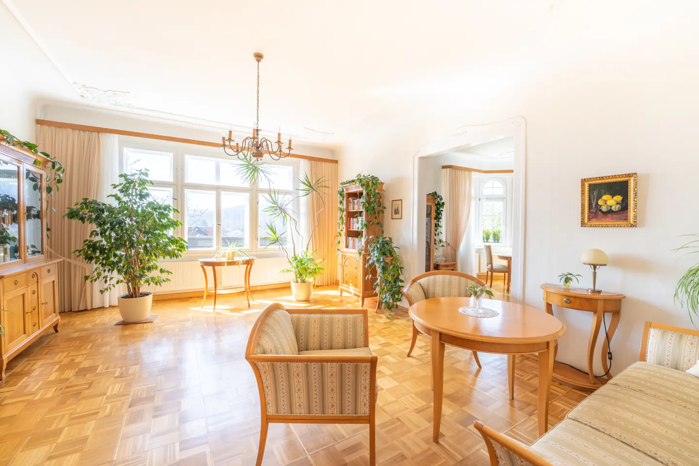 Bright living room with parquet floors, wooden furniture, striped armchairs, a round dining table, and several houseplants.