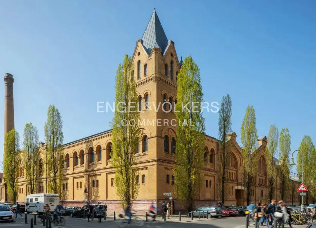 Historic yellow brick building with a tall tower, arched windows, and a chimney, surrounded by trees under a blue sky.