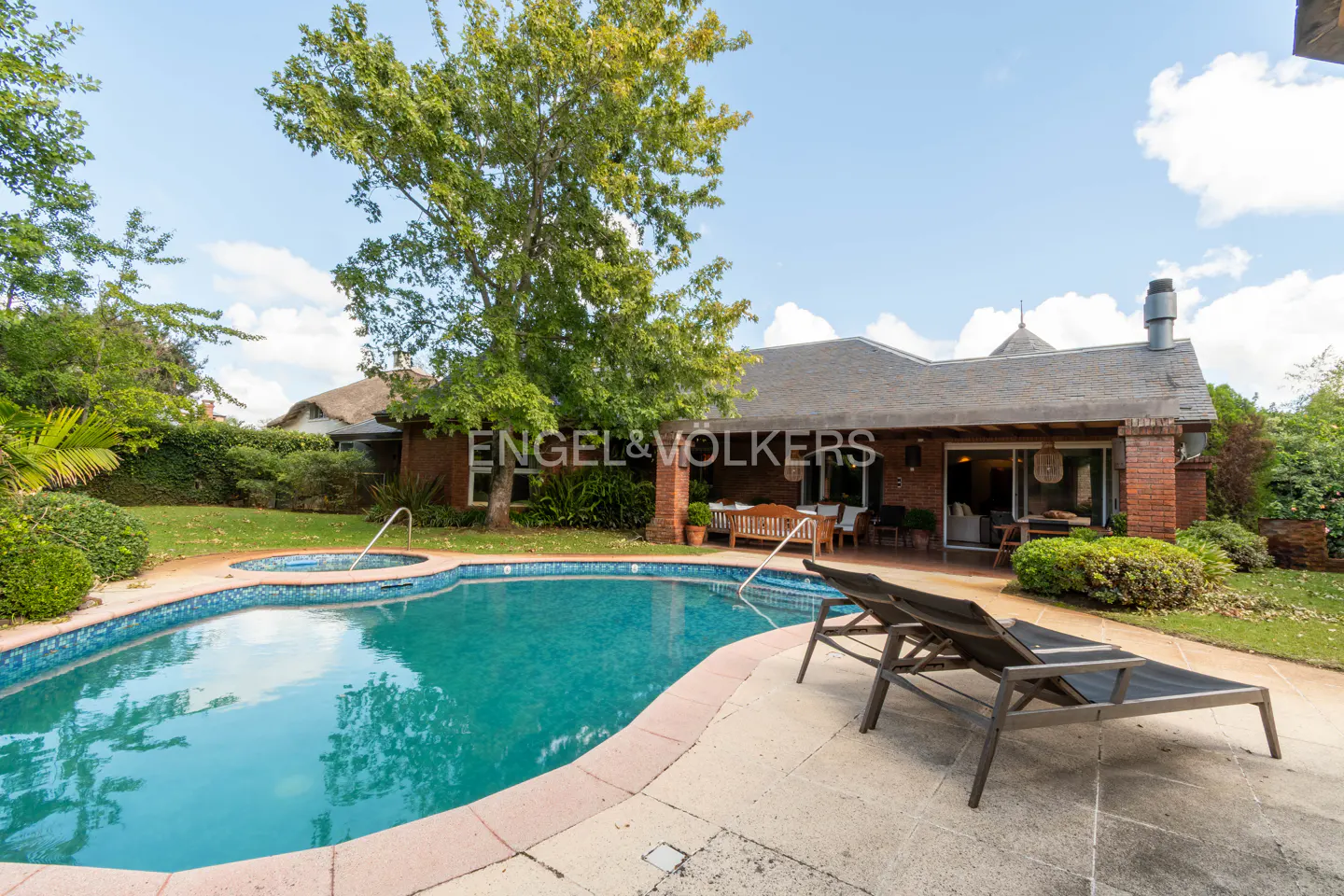 A backyard with a pool, two lounge chairs, and a brick house with a chimney under a blue sky.