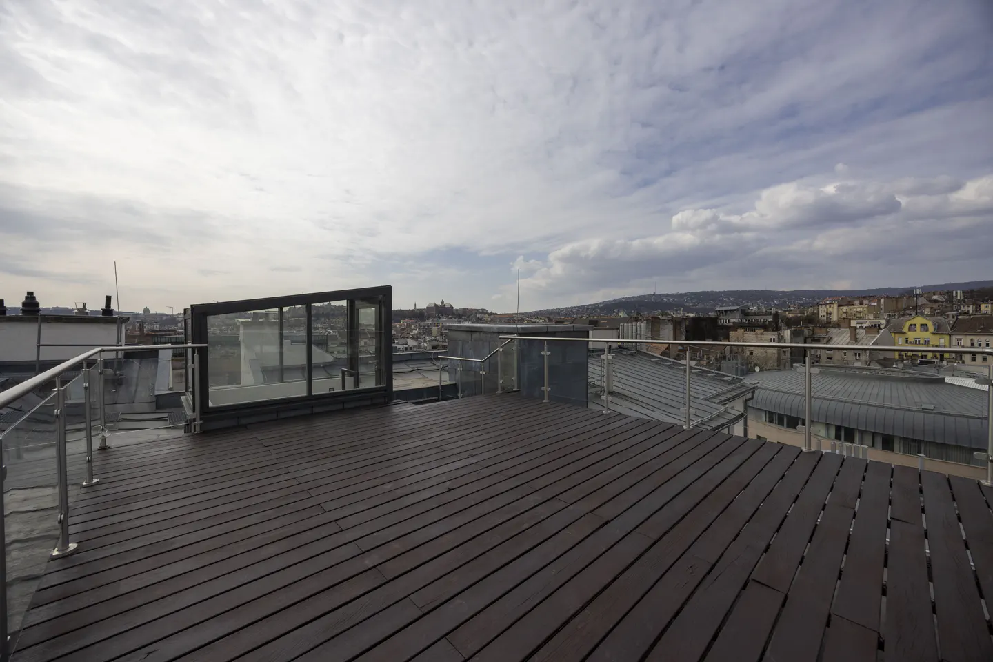 Rooftop deck with dark wood planks, glass railings, and a city view under a cloudy sky.