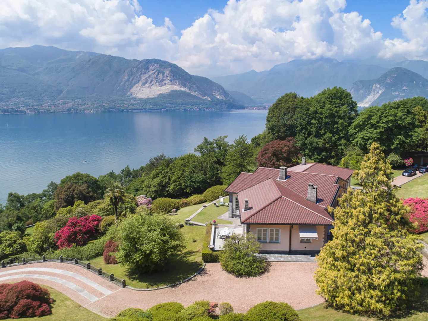 Aerial view of a beige house with a red tile roof, surrounded by green trees and colorful flowers, overlooking a blue lake and mountains.