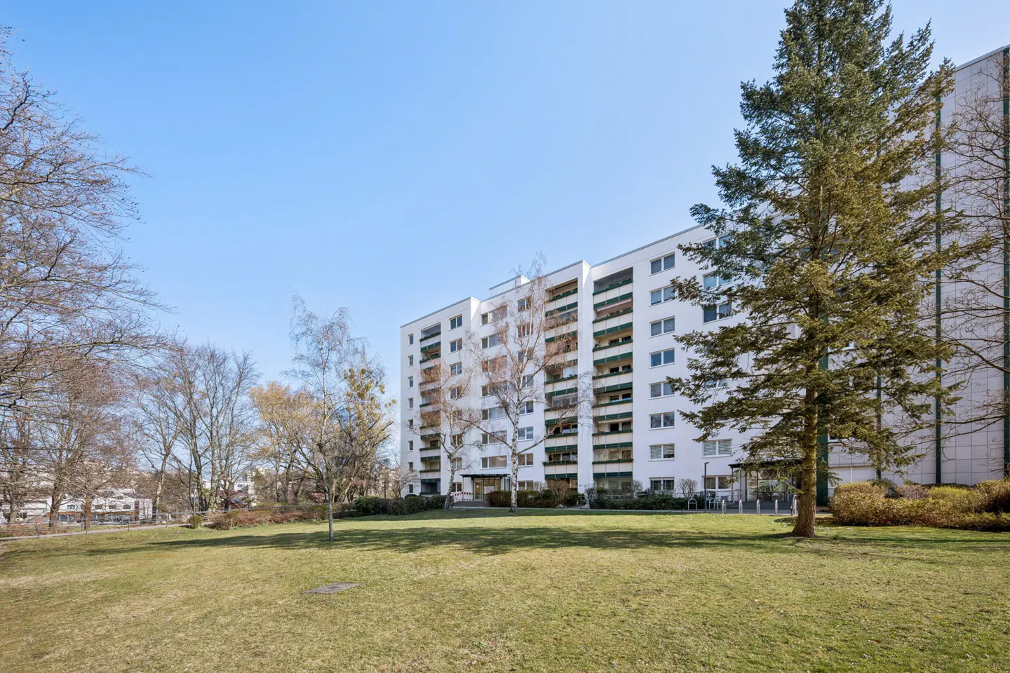 Exterior view of a white apartment building with balconies, surrounded by green lawn and trees under a blue sky.