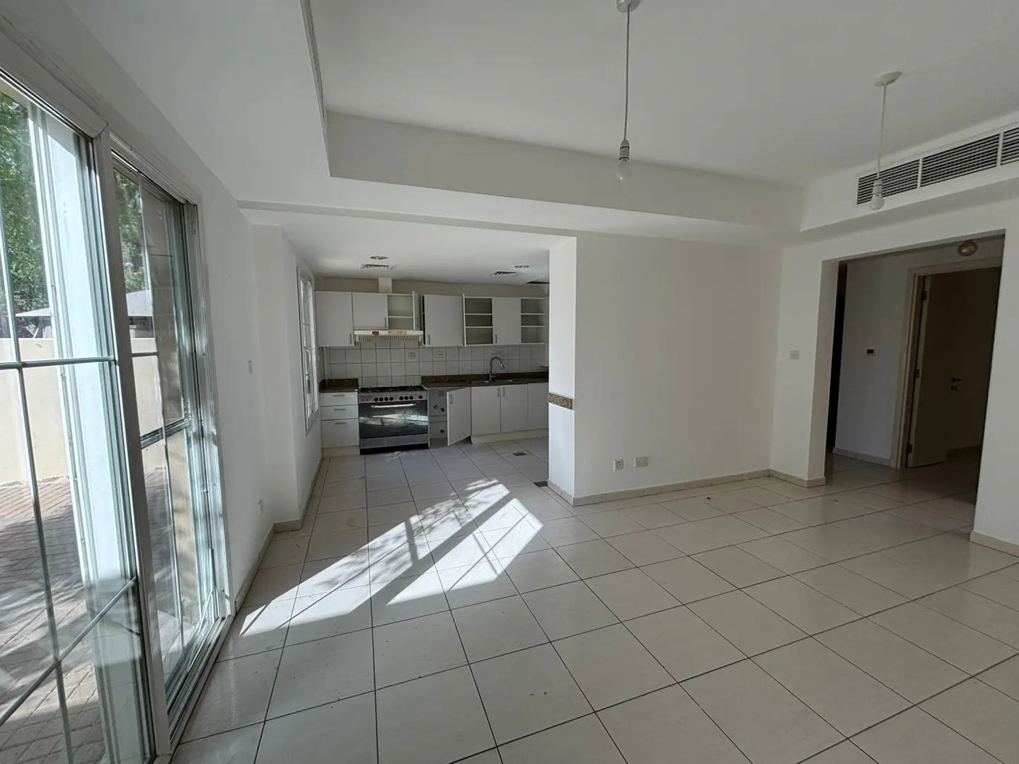 Bright, empty room with white walls and tile floor. Kitchen with white cabinets visible in the background. Large sliding glass doors to the left.