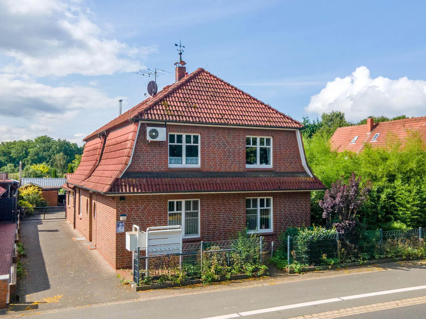 Two-story brick house with a red tile roof, white windows, and a small fenced yard on a sunny day.