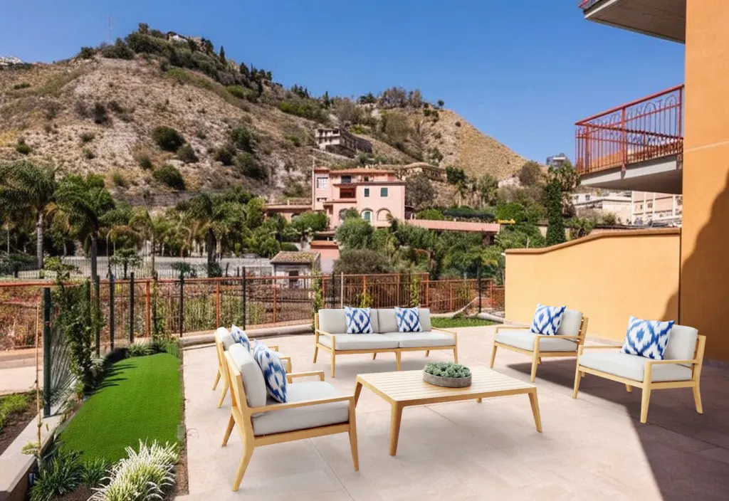 Outdoor patio with light wood furniture, white cushions, and blue patterned pillows, overlooking a hillside and villa.