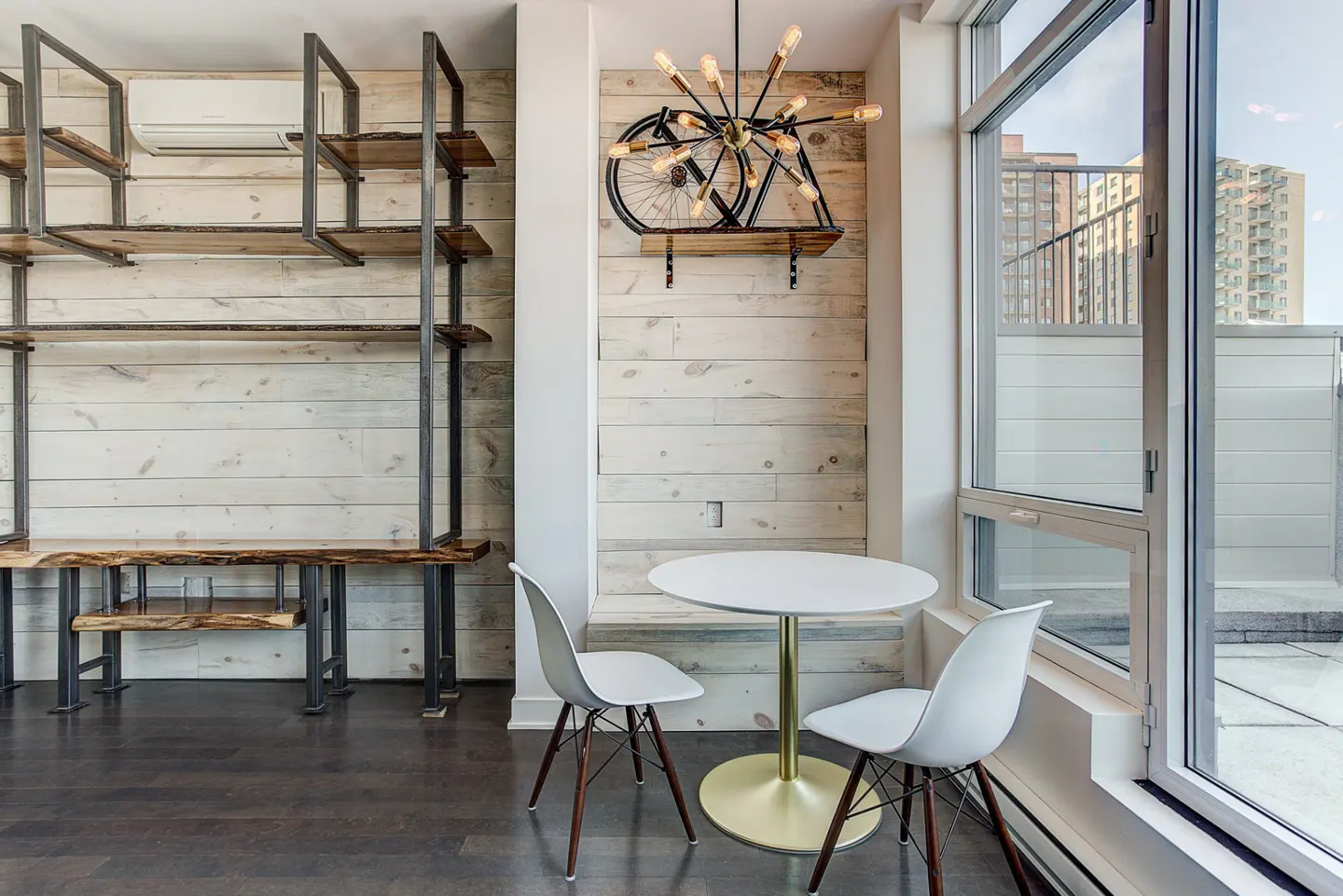 A modern apartment interior with a white table and chairs near a large window. A bicycle-shaped light fixture hangs on a white wood-paneled wall.