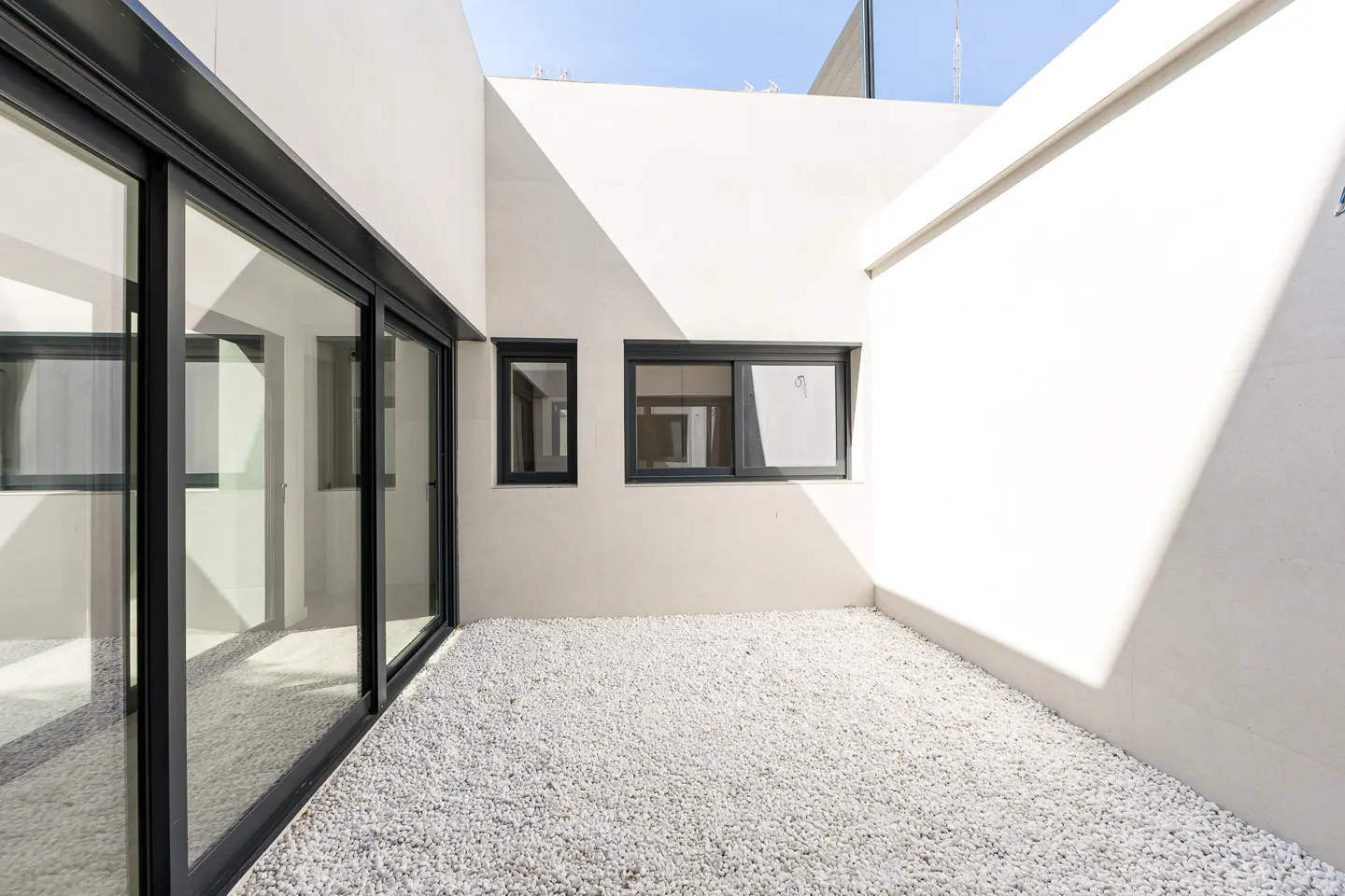 Minimalist courtyard with white walls, black-framed windows, and white gravel ground cover. Sunlight creates shadows on the walls.