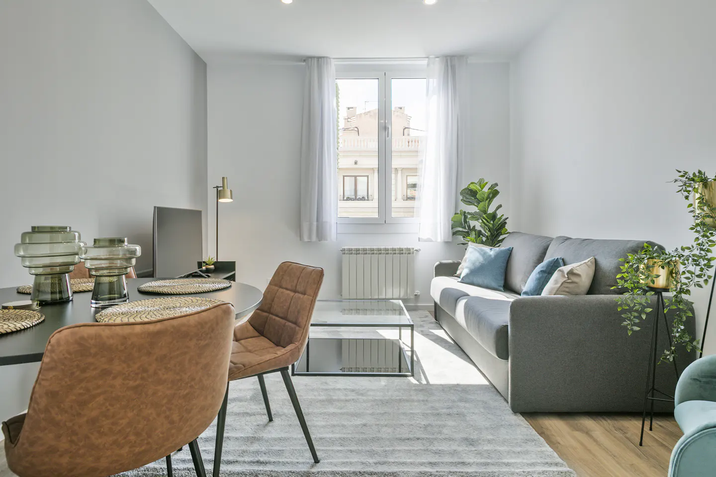 Bright, modern living room with a gray sofa, brown chairs, and a round black table. Plants add a touch of nature. Sunlight streams through the window.