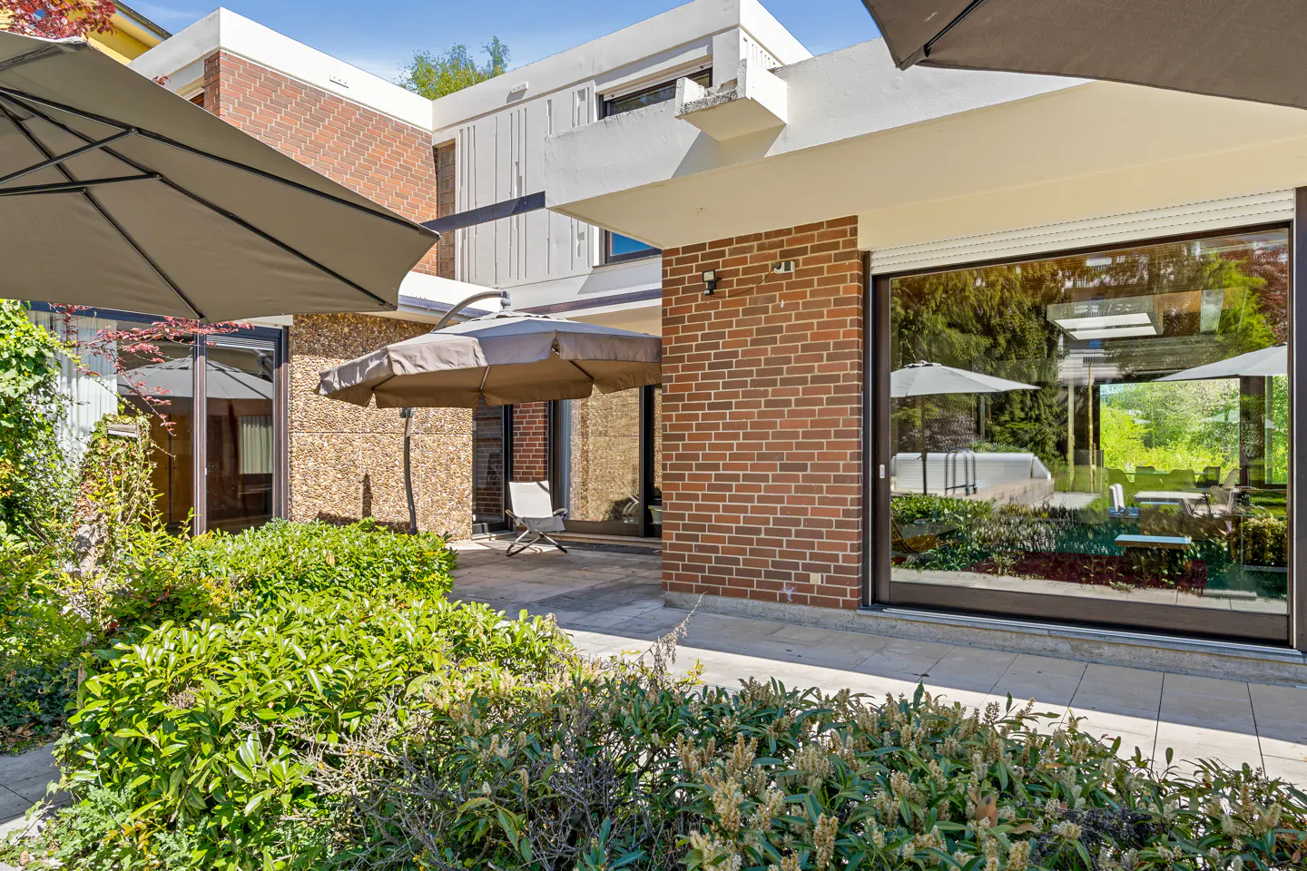 Modern home exterior with brick and stone walls, large windows, patio umbrellas, and lush green landscaping.