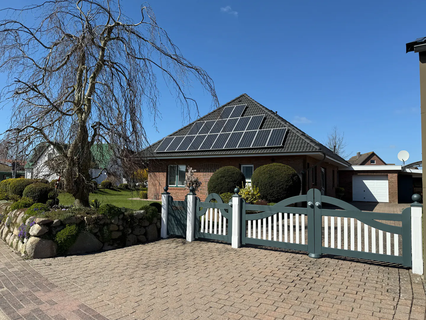 A brick house with solar panels on the roof, a gray and white gate, and a tree with bare branches.