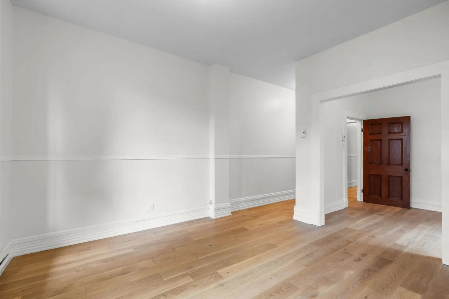 Bright, empty room with light wood floors, white walls, and a dark wood door visible through an open doorway.