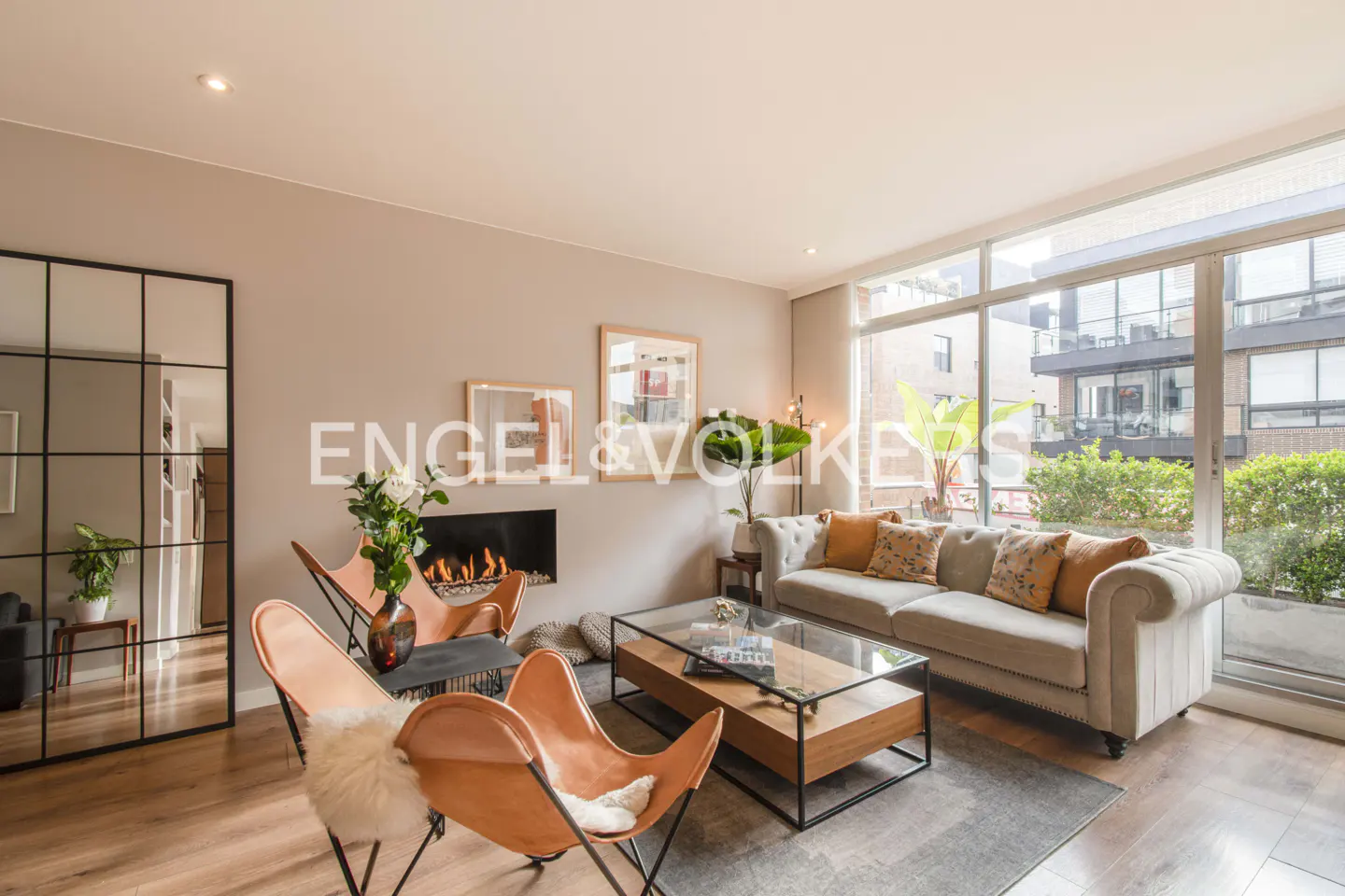 Living room with hardwood floors, a gray sofa, and two orange butterfly chairs. A fireplace and large windows add warmth and light.