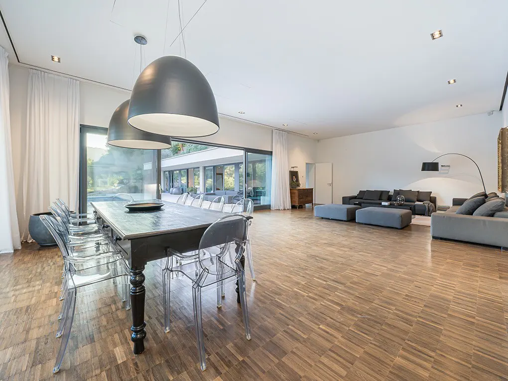 Bright, open-plan living and dining area with wood floors, gray sofas, and a long dark wood table with clear chairs. Large windows offer outdoor views.