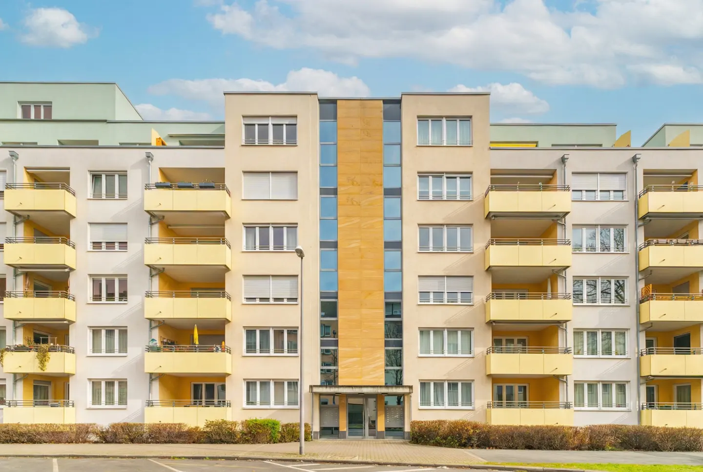 Exterior view of a multi-story apartment building with yellow balconies and a central glass facade under a blue sky.