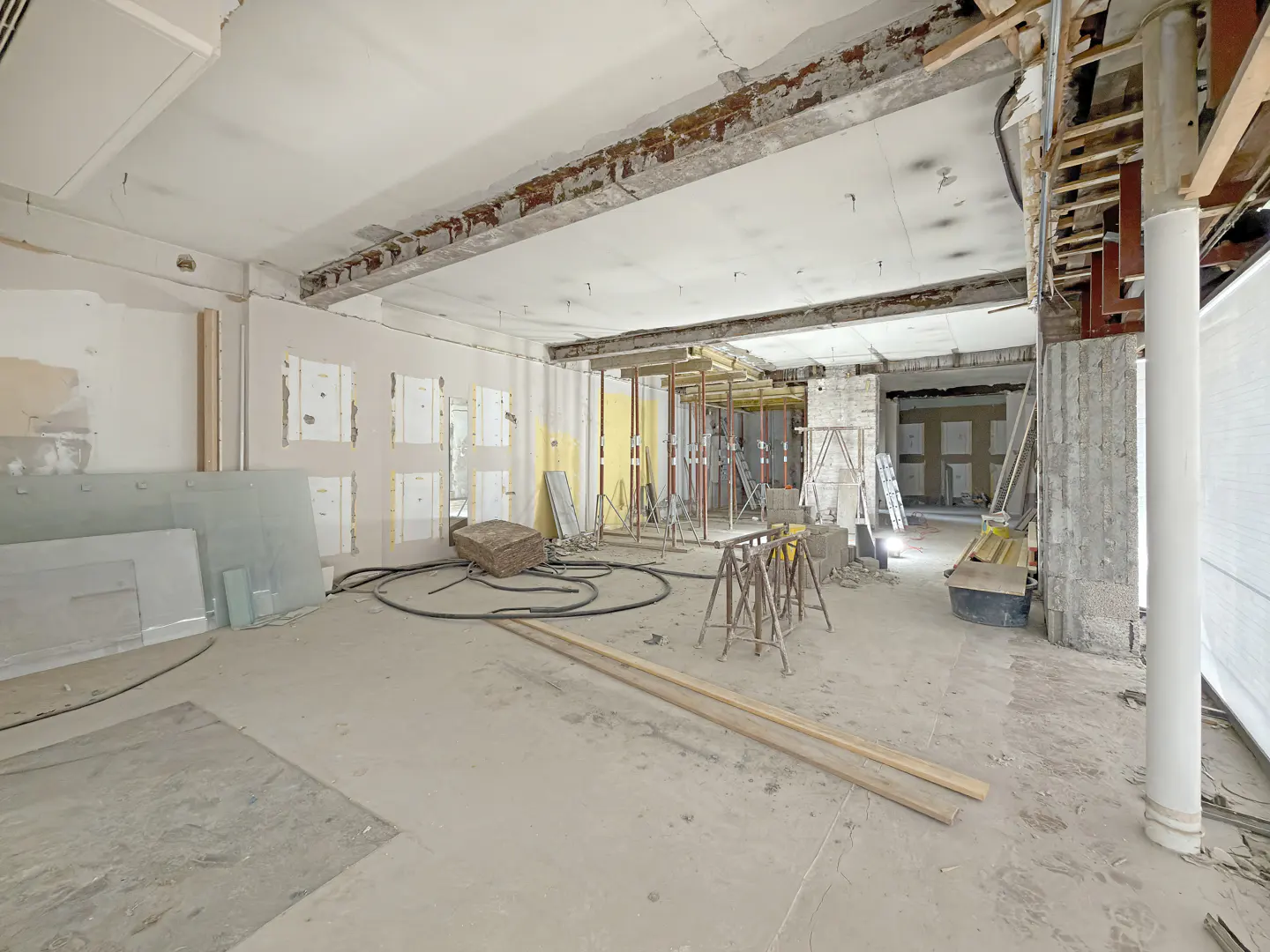 Interior view of a commercial space undergoing renovation, with exposed beams, scaffolding, and construction materials scattered throughout.