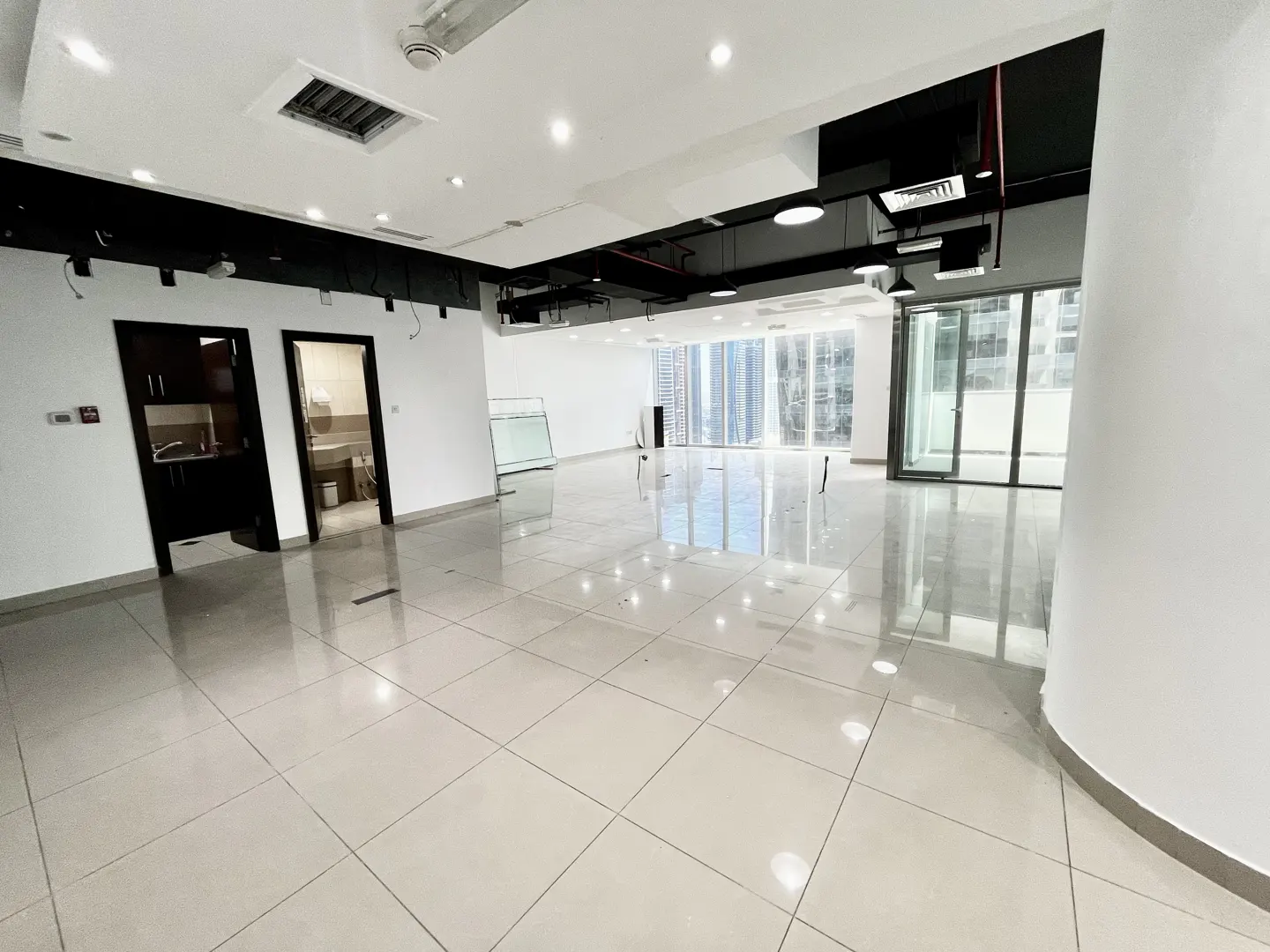 Bright, empty office space with white tile floors, black ceiling fixtures, and large windows with a city view. Two doorways lead to a kitchen and bathroom.