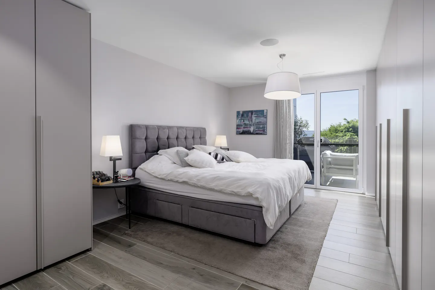 Modern bedroom with a grey tufted bed, white bedding, built-in wardrobes, and a balcony door with a view of greenery.