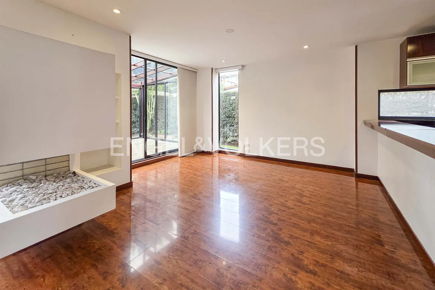 Bright, empty living room with hardwood floors, white walls, and a modern fireplace filled with white stones. Large windows overlook a green garden.