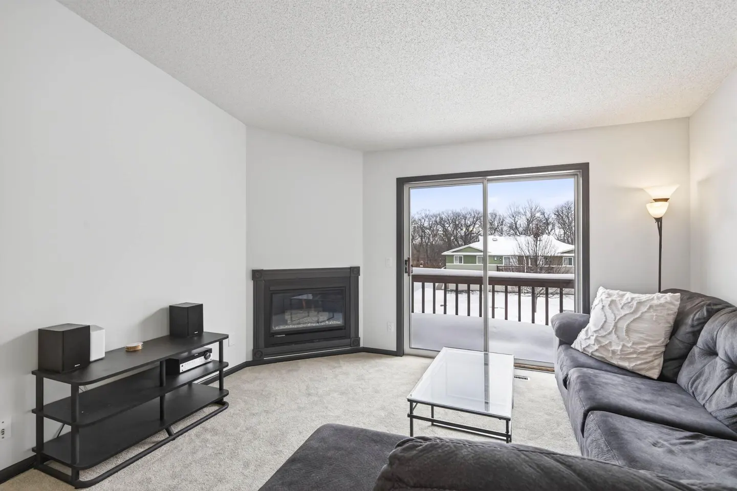 Living room with gray sofa, glass table, and black fireplace. Sliding glass doors lead to a snow-covered deck. A black TV stand sits against a white wall.