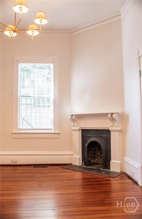 Empty room with hardwood floors, a white fireplace, and a window with blinds. A gold chandelier hangs from the ceiling.