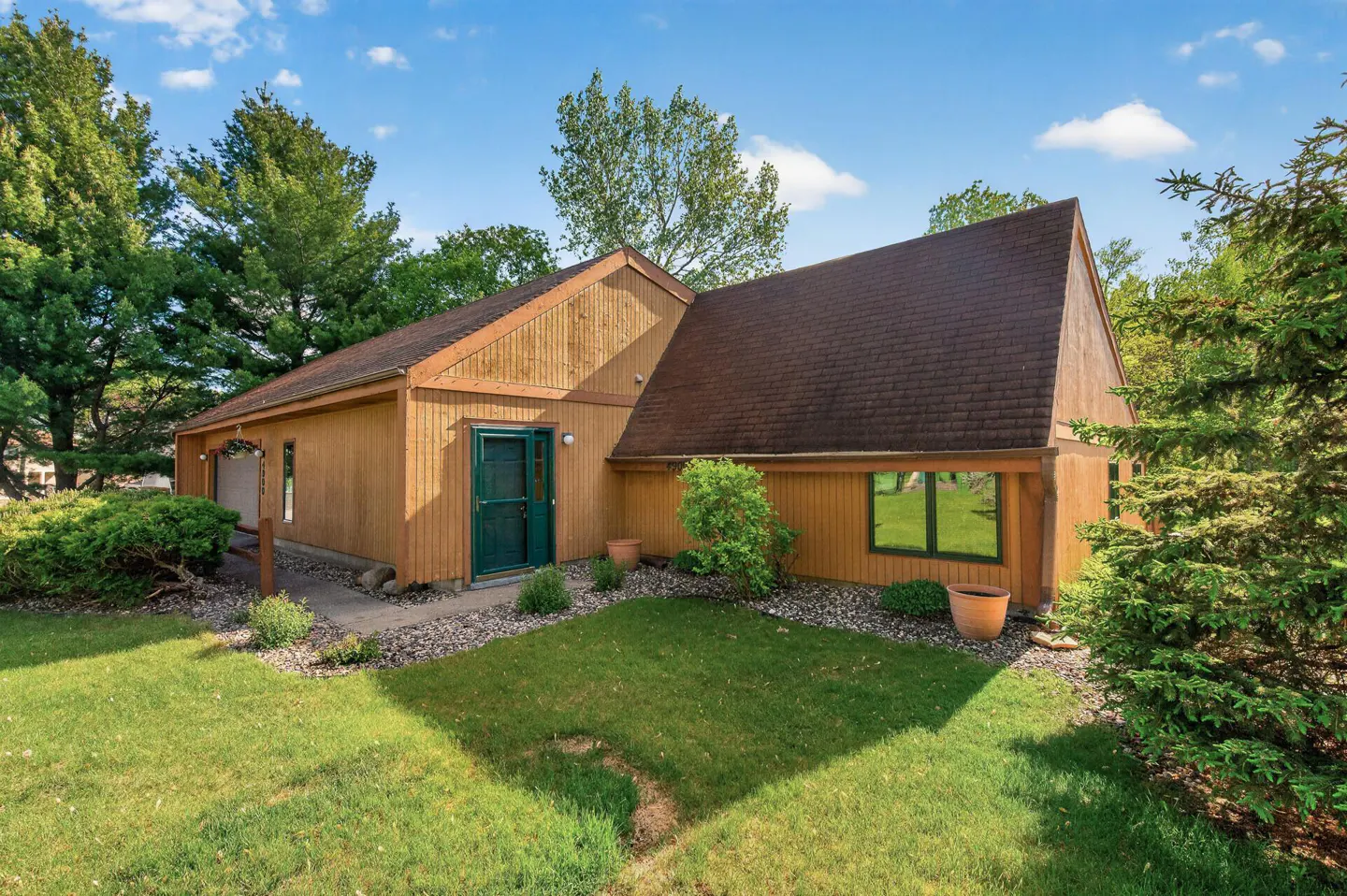 Exterior view of a tan house with a brown roof, green door, and green-trimmed windows, surrounded by green grass and trees.