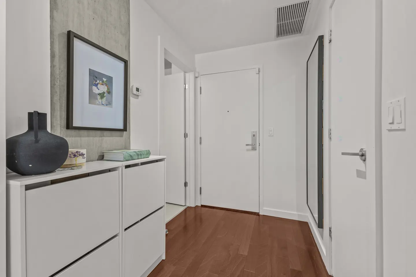 Hallway with dark wood floors, white walls, and white shoe cabinet topped with a black vase and framed art.