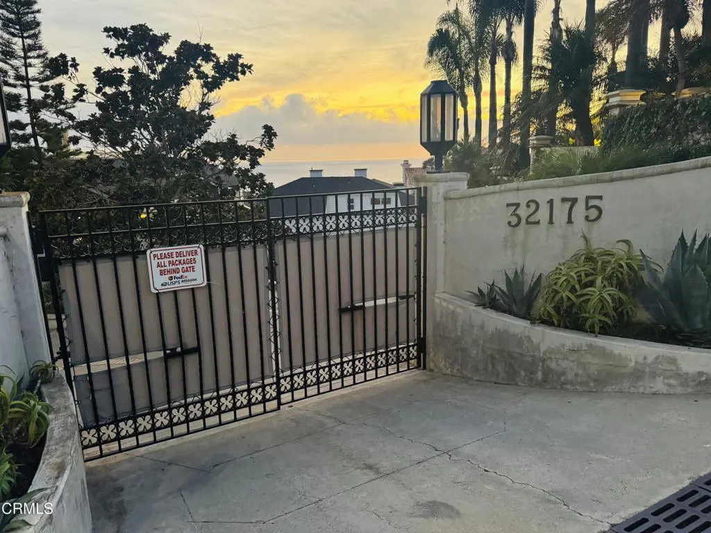 Gated driveway entrance to a home with address 32175 on a stucco wall. Ocean view and sunset in the background.