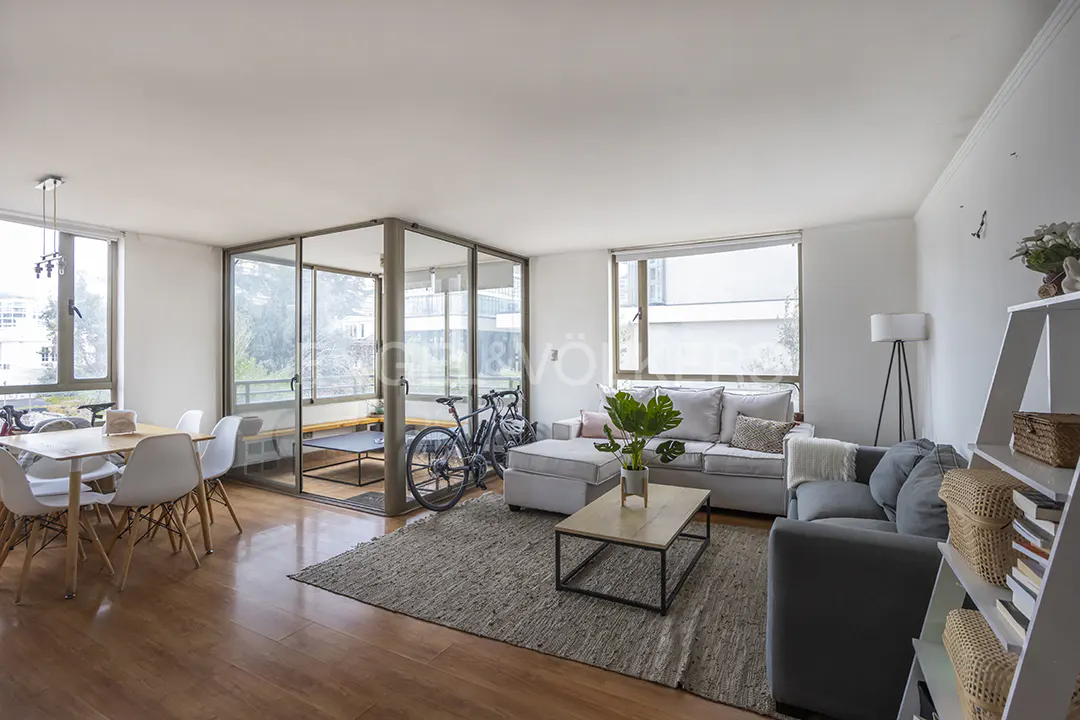 Bright, open living space with wood floors, white walls, and large windows. A dining table, sofas, a bike, and a bookshelf are visible.