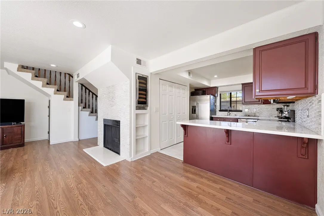 A bright, open-concept living space with wood floors, a fireplace, and a kitchen island with maroon cabinets. Stairs lead to the upper level.